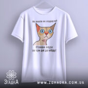 Футболка як швидко схуднути. Біла футболка, підвішена на вішалці, з принтом кішки та текстом: 'Не знаєте як схуднути? Стакан води за три дні до обіду!' Фон світлий, без зайвих деталей.