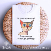 Футболка як швидко схуднути. Біла футболка, складена на плетеній кошику, з принтом кішки та текстом: 'Не знаєте як схуднути? Стакан води за три дні до обіду!' Фон натуральний, що додає затишку.