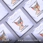 Футболка як швидко схуднути. Біла футболка з принтом, на якому зображена кішка з великими блакитними очима та червоним носом. Принт містить текст: 'Не знаєте як схуднути? Стакан води за три дні до обіду!' Фон нейтральний, без додаткових елементів.