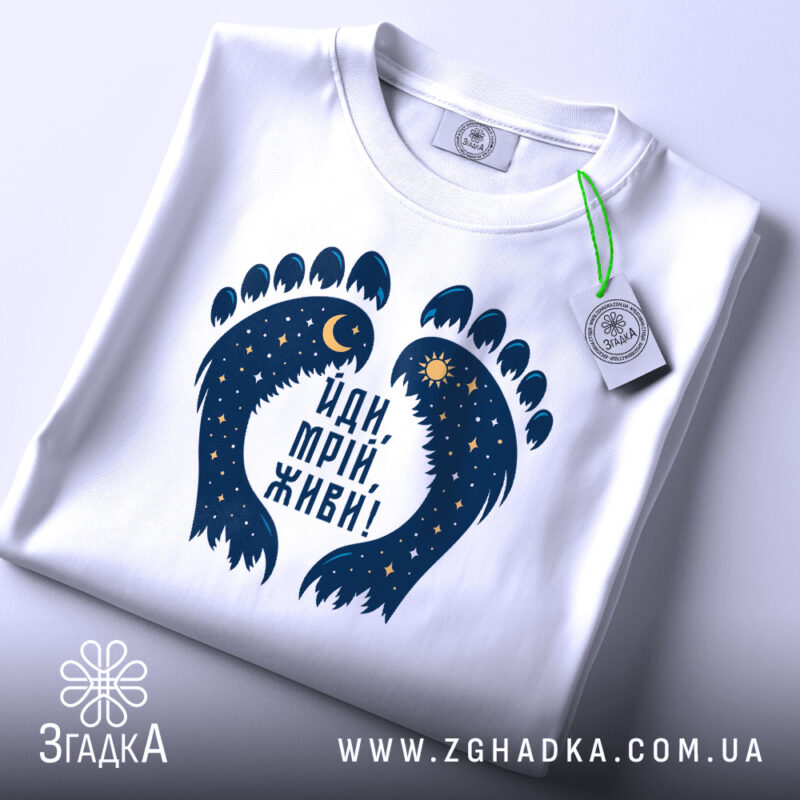 Футболка йди мрій живи, біла, зображена зверху, фон світло-сірий, принт у вигляді слідів з зірками та місяцем, на комірці є етикетка.