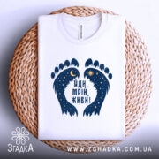 Футболка йди мрій живи, біла, зображена на плоскій поверхні, фон світлий, принт у вигляді слідів з зірками та місяцем, розташована на плетеній основі.