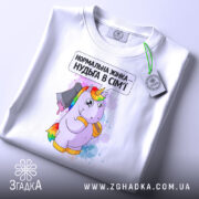 Футболка єдинорог нормальна жінка, біла, складена на столі, фон світло-сірий, принт з єдинорогом, що тримає ніж, текст зверху: 'Нормальна жінка – нудьга в сім'ї', на футболці зелена етикетка.