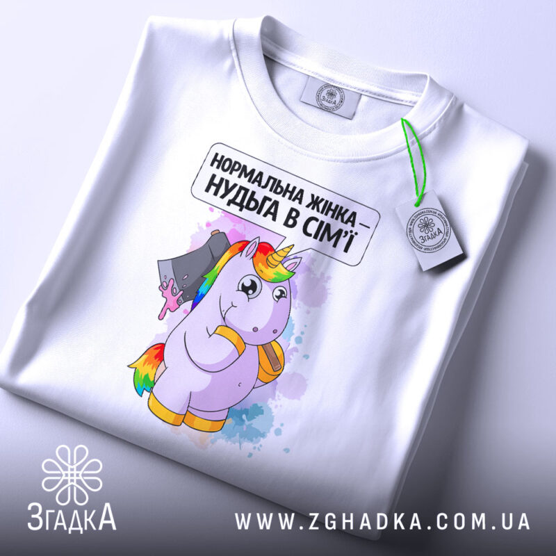 Футболка єдинорог нормальна жінка, біла, складена на столі, фон світло-сірий, принт з єдинорогом, що тримає ніж, текст зверху: 'Нормальна жінка – нудьга в сім'ї', на футболці зелена етикетка.