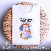 Футболка єдинорог нормальна жінка, біла, складена на плетеному кошику, фон світло-сірий, принт з єдинорогом, що тримає ніж, текст зверху: 'Нормальна жінка – нудьга в сім'ї'.