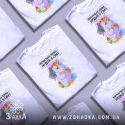 Футболка єдинорог нормальна жінка, біла, зображена на манекені, фон світло-сірий, принт з веселим єдинорогом, який тримає ножа з рожевою рідиною, текст зверху: 'Нормальна жінка – нудьга в сім'ї'.