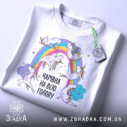 Футболка єдинороги білого кольору, складена на поверхні, фон світло-сірий, принт з веселкою та єдинорогами, текст "ЧАРІВНА НА ВСЮ ГОЛОВУ" в чорному кольорі, на футболці є етикетка.