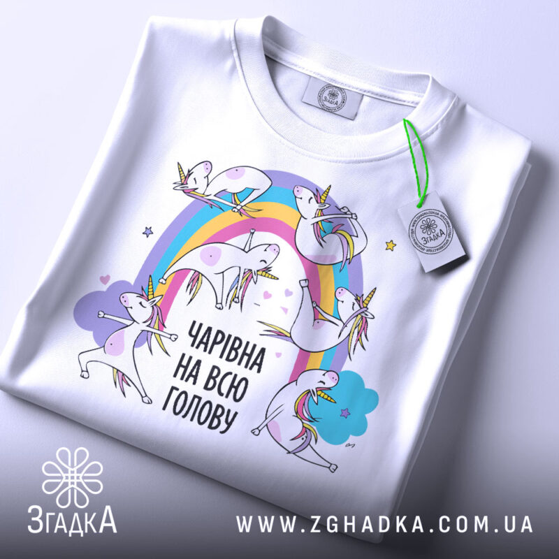 Футболка єдинороги білого кольору, складена на поверхні, фон світло-сірий, принт з веселкою та єдинорогами, текст "ЧАРІВНА НА ВСЮ ГОЛОВУ" в чорному кольорі, на футболці є етикетка.