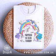 Футболка єдинороги білого кольору, складена на плетеній кошику, фон світло-сірий, принт з веселкою та єдинорогами, текст "ЧАРІВНА НА ВСЮ ГОЛОВУ" в чорному кольорі.