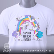 Футболка єдинороги білого кольору, складена на поверхні, фон світло-сірий, принт з веселкою та єдинорогами, текст "ЧАРІВНА НА ВСЮ ГОЛОВУ" в чорному кольорі, на футболці є етикетка.