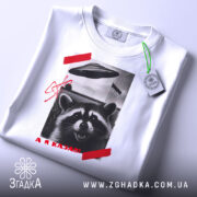 Футболка єнот гумор НЛО білого кольору, зображена зверху, на фоні світлого кольору, принт з єнотом, літаючою тарілкою та червоною смужкою, на комірці видна етикетка.