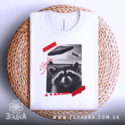 Футболка єнот гумор НЛО білого кольору, зображена зверху на плетеній кошику, фон світлий, принт з єнотом, літаючою тарілкою та червоною смужкою, видна етикетка на комірці.