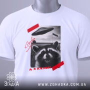 Футболка єнот гумор НЛО білого кольору, зображена зверху, на фоні світлого кольору, принт з єнотом, літаючою тарілкою та червоною смужкою, на комірці видна етикетка.