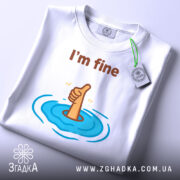 Футболка I'm fine білого кольору, зображена з верхнього ракурсу, складена на поверхні, фон світло-сірий, принт з рукою, що піднімає великий палець, оточений блакитними хвилями, з етикеткою на зеленому шнурку.