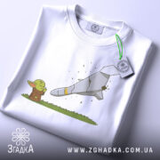 Футболка йода зупиняє шахед, біла футболка, складена на столі, з принтом йоди та ракети, фон світло-сірий, деталі принта чіткі, з етикеткою на зеленому шнурку.