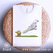 Футболка йода зупиняє шахед, біла футболка, складена на плетеному кошику, з принтом йоди та ракети, фон світло-сірий, деталі принта яскраві, з зеленими та сірими кольорами.