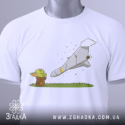 Футболка йода зупиняє шахед, біла футболка, складена на столі, з принтом йоди та ракети, фон світло-сірий, деталі принта чіткі, з етикеткою на зеленому шнурку.