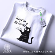 Футболка їсти чи не їсти. Біла футболка складена на столі, з принтом чорного кота, який тримає круасан. На футболці є етикетка з зеленим шнурком. Фон світло-сірий.