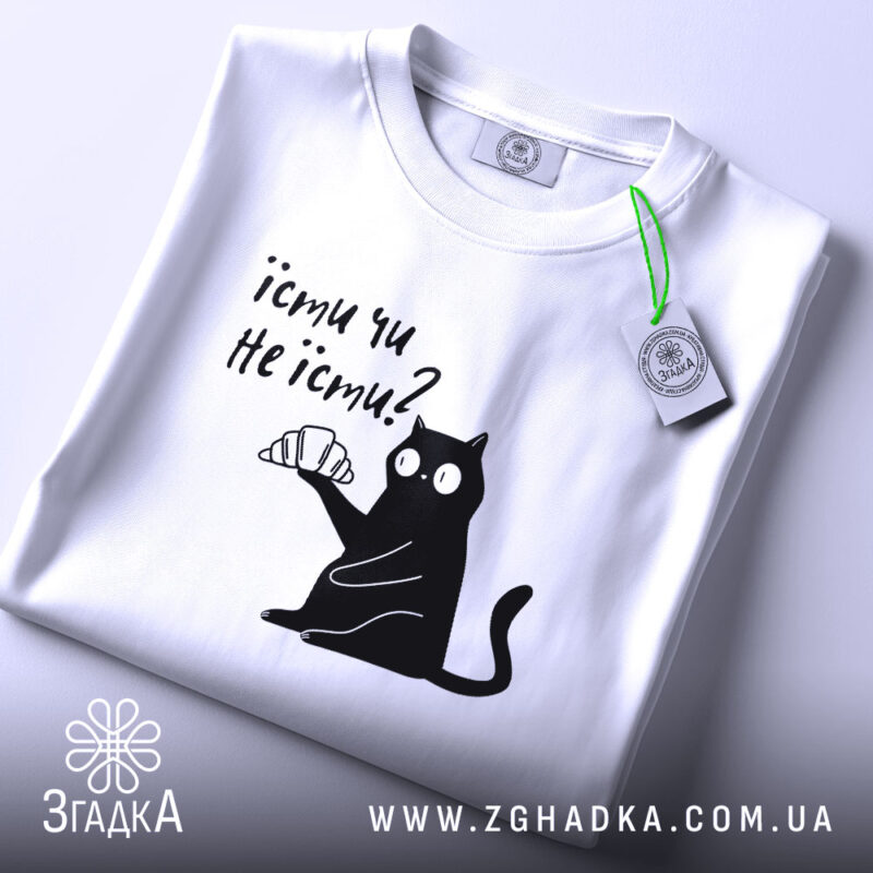 Футболка їсти чи не їсти. Біла футболка складена на столі, з принтом чорного кота, який тримає круасан. На футболці є етикетка з зеленим шнурком. Фон світло-сірий.