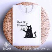 Футболка їсти чи не їсти. Біла футболка складена на плетеному кошику, з принтом чорного кота, який тримає круасан. Фон світло-сірий, видно частину етикетки.