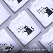 Футболка їсти чи не їсти. Біла футболка на манекені, з принтом чорного кота, який тримає круасан. Фон світло-сірий, футболка має короткі рукави.