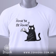 Футболка їсти чи не їсти. Біла футболка складена на столі, з принтом чорного кота, який тримає круасан. На футболці є етикетка з зеленим шнурком. Фон світло-сірий.