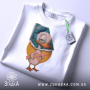 Футболка качур боєць білого кольору, складена на столі, фон світло-сірий, принт з качуром у капелюсі, що тримає гвинтівку, з етикеткою на рукаві.