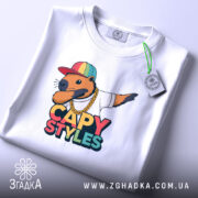 Футболка капібара білого кольору, складена на столі, з принтом капібари в кепці та золотому ланцюгу, на етикетці зелена стрічка, фон світло-сірий.