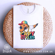 Футболка капібара білого кольору, складена на плетеній кошику, з принтом капібари в кепці та золотому ланцюгу, фон світло-сірий.