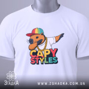 Футболка капібара білого кольору, складена на столі, з принтом капібари в кепці та золотому ланцюгу, на етикетці зелена стрічка, фон світло-сірий.