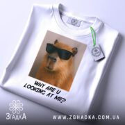 Футболка капібара з жартом, біла, складена на столі, фон світлий, принт з капібарами в окулярах та текстом "WHY ARE U LOOKING AT ME?" під ним, з етикеткою на рукаві.