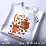 Футболка кавовий характер, біла, зображена зверху, фон світлий, принт з чашкою кави, кавомолкою та написом 'makes me nicer', з етикеткою на рукаві.