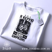 Футболка керую цим цирком, білого кольору, зображена зверху, на ній видно етикетку з зеленим шнурком, фон світло-сірий, принт з тигром у капелюсі та текстом "Я КЕРУЮ ЦИРКОМ!" у чорному кольорі.