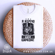 Футболка керую цим цирком, білого кольору, зображена зверху на плетеній основі, фон білий, принт з тигром у капелюсі та текстом "Я КЕРУЮ ЦИРКОМ!" у чорному кольорі.