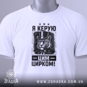 Футболка керую цим цирком, білого кольору, зображена зверху, на ній видно етикетку з зеленим шнурком, фон світло-сірий, принт з тигром у капелюсі та текстом "Я КЕРУЮ ЦИРКОМ!" у чорному кольорі.