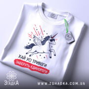 Футболка хай тривоги, біла, зображена зверху, на столі, фон білий, принт з єдинорогом у сонцезахисних окулярах, з крилами, з зеленим ярликом.