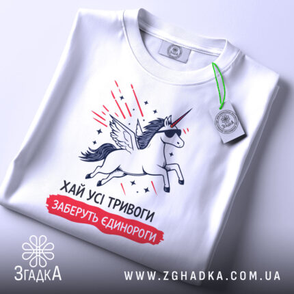Футболка хай тривоги, біла, зображена зверху, на столі, фон білий, принт з єдинорогом у сонцезахисних окулярах, з крилами, з зеленим ярликом.