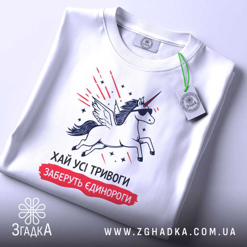 Футболка хай тривоги, біла, зображена зверху, на столі, фон білий, принт з єдинорогом у сонцезахисних окулярах, з крилами, з зеленим ярликом.