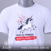 Футболка хай тривоги, біла, зображена зверху, на столі, фон білий, принт з єдинорогом у сонцезахисних окулярах, з крилами, з зеленим ярликом.