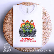 Футболка хардкор без компромісів, біла, складена на плетеній кошику, фон світлий, принт з зеленим персонажем, що тримає гантелі, з червоним написом 'ХАРДКОР БЕЗ КОМПРОМІСІВ'.
