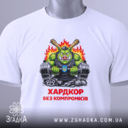 Футболка хардкор без компромісів, біла, складена на поверхні, фон світлий, принт з зеленим персонажем у чорному майці, що тримає гантелі, з червоним написом 'ХАРДКОР БЕЗ КОМПРОМІСІВ'.