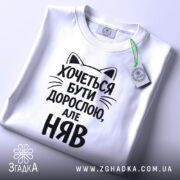 Футболка хочеться бути дорослою. Біла футболка з чорним принтом, зображена зверху на плоскій поверхні. Принт містить текст з котячими вушками, що говорить: 'Хочеться бути дорослою, але НЯВ', поряд з етикеткою.