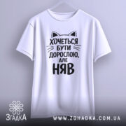 Футболка хочеться бути дорослою. Біла футболка з чорним принтом, зображена з фронтального ракурсу на манекені. Принт містить текст з котячими вушками, що говорить: 'Хочеться бути дорослою, але НЯВ'.