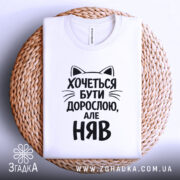 Футболка хочеться бути дорослою. Біла футболка з чорним принтом, зображена зверху на плоскій поверхні, розташована на плетеній основі. Принт містить текст з котячими вушками, що говорить: 'Хочеться бути дорослою, але НЯВ'.
