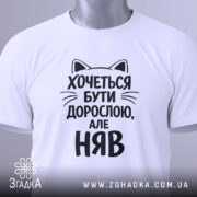 Футболка хочеться бути дорослою. Біла футболка з чорним принтом, зображена зверху на плоскій поверхні. Принт містить текст з котячими вушками, що говорить: 'Хочеться бути дорослою, але НЯВ', поряд з етикеткою.