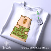 Футболка хочу обіймати, біла, зображена зверху, фон світло-сірий, принт з ведмедем, який тримає зелену стрічку з написом 'Олі потрібно обіймати', на футболці є етикетка з зеленим шнурком.