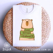 Футболка хочу обіймати, біла, зображена зверху на плетеній основі, фон білий, принт з ведмедем, який тримає зелену стрічку з написом 'Олі потрібно обіймати'.