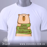 Футболка хочу обіймати, біла, зображена зверху, фон світло-сірий, принт з ведмедем, який тримає зелену стрічку з написом 'Олі потрібно обіймати', на футболці є етикетка з зеленим шнурком.