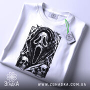 Футболка хорор скрім, складена біла футболка з принтом чорного кольору, що зображує маску з фільму жахів, на фоні світло-сірого кольору, з етикеткою зеленого кольору, що звисає з коміра.