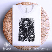 Футболка хорор скрім, складена біла футболка на плетеній кошику, з принтом чорного кольору, що зображує маску з фільму жахів, на фоні світло-сірого кольору, деталі принта включають черепи та елементи, що нагадують про жахи.