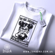 Футболка хто тут головний. Біла футболка, складена на поверхні, з принтом чорного кольору. На футболці видно персонажа з окулярами, який вказує пальцем. На футболці є етикетка з зеленим шнурком. Фон світло-сірий.